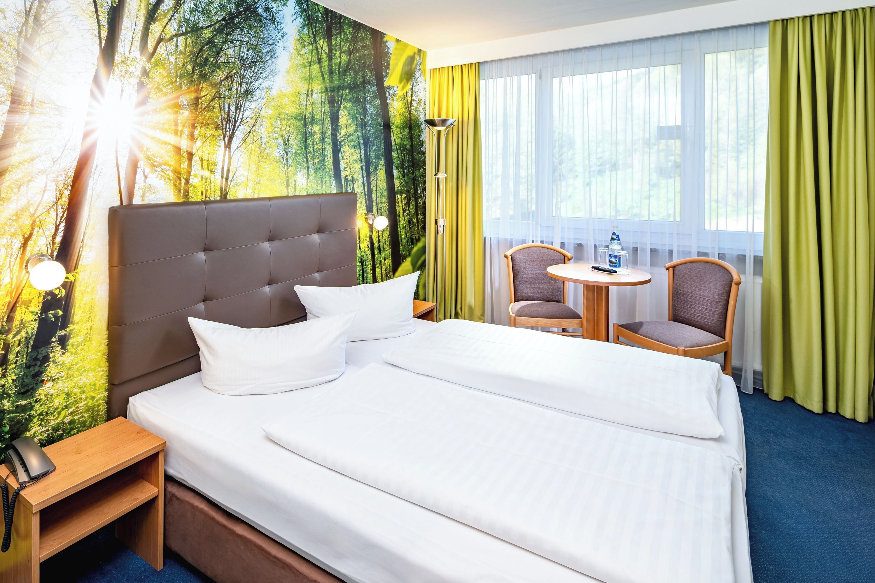 A hotel room with a large bed, a bedside table with a phone, a small round table with chairs, and a window with curtains.