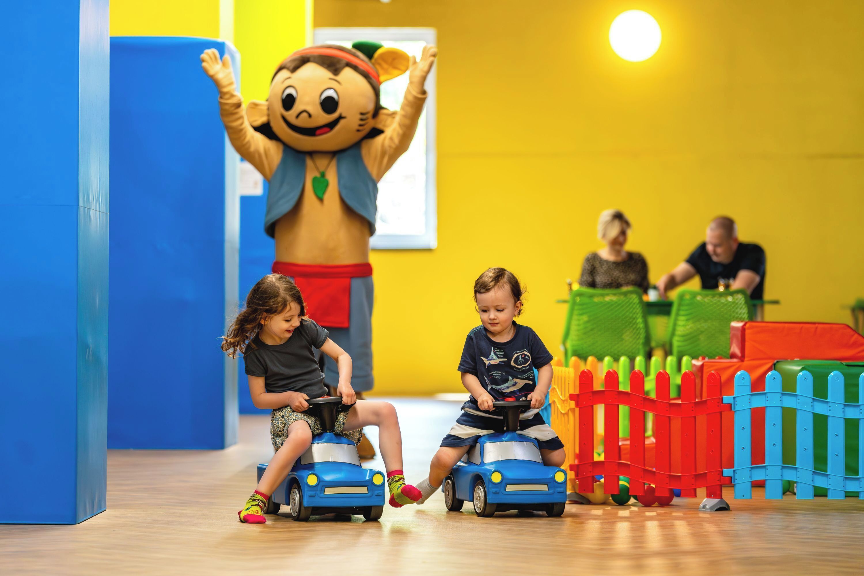 Children playing with toy cars in a brightly colored room with a large mascot figure.