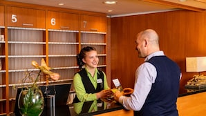 Een hotel receptie met houten lambrisering, nummerde kamers, een receptiebalie en een vaas met een groen blad.