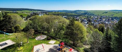 Un parque infantil con diversas estructuras de juego, rodeado de árboles y vistas a un pueblo al fondo.