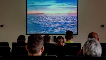 A group of people seated in a row watching a large screen displaying a sunset over the ocean.