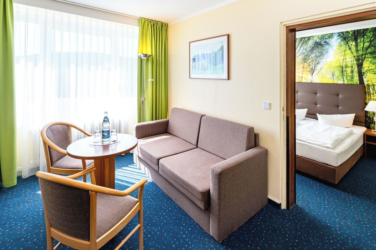 Une chambre d’hôtel avec un canapé, deux fauteuils, une petite table avec une bouteille d’eau, un lit et une fenêtre avec des rideaux.