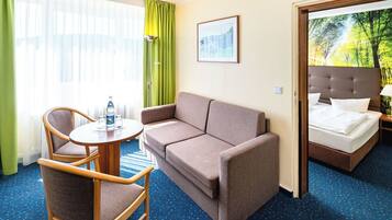 A hotel room with a sofa, two chairs, a small table with a water bottle, a bed, and a window with curtains.