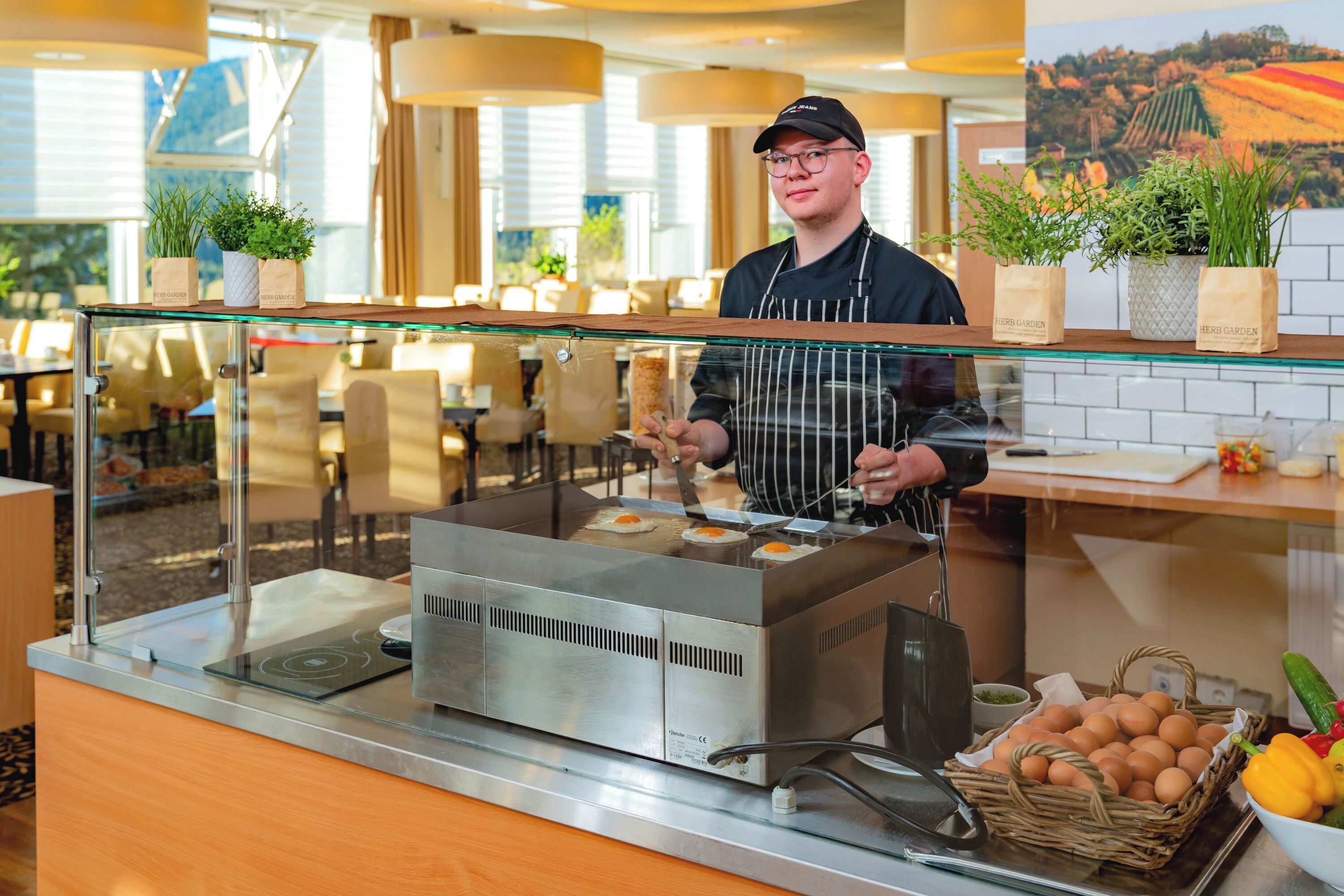 A kitchen area with a food preparation station, including a fryer and a display case with food items. There are potted plants on the counter, and a basket of eggs and vegetables is visible.