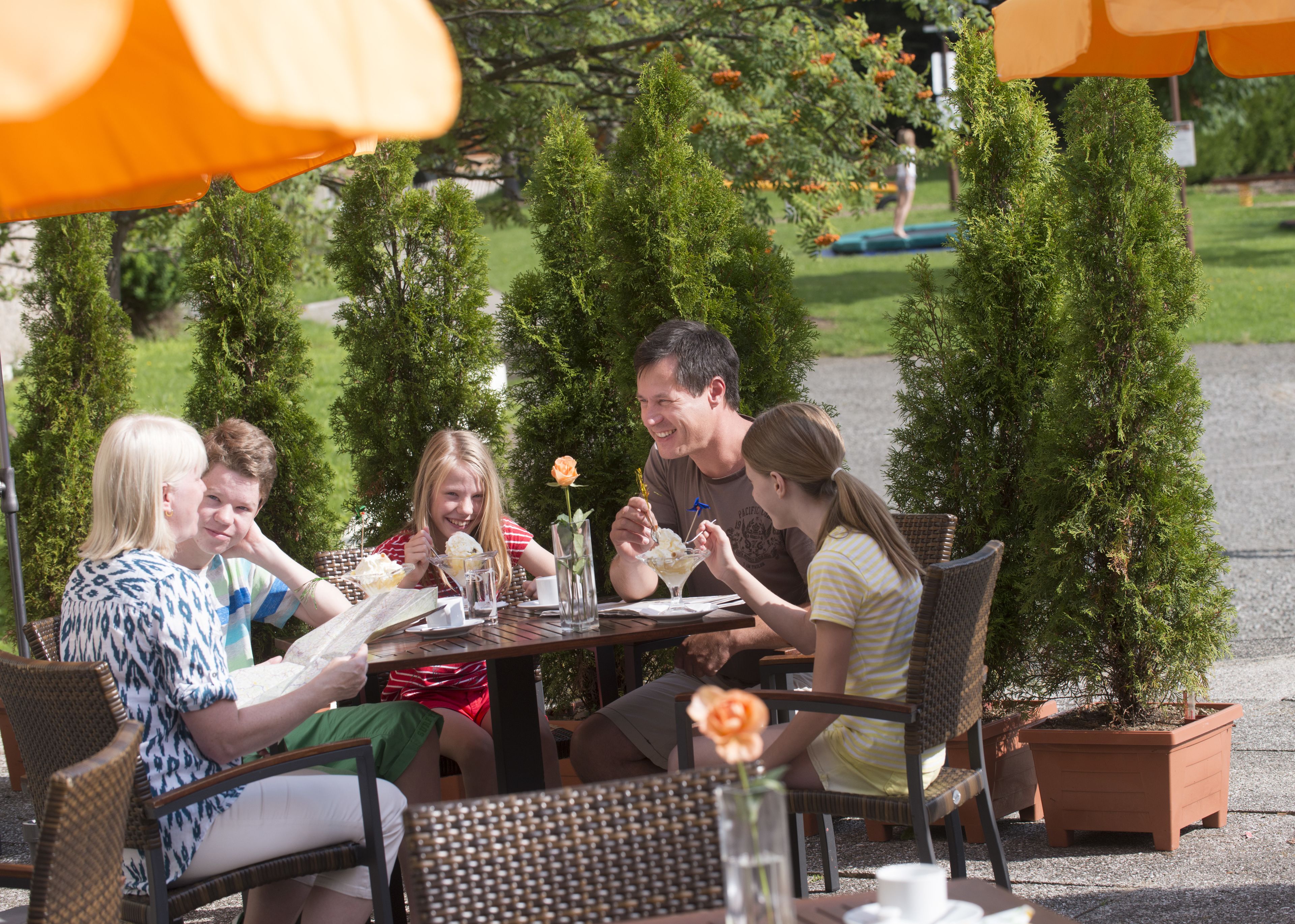 A group of people dining at an outdoor table with potted plants and orange umbrellas.
