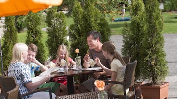 Un grupo de personas cenando en una mesa al aire libre, con plantas en macetas y sombrillas naranjas.