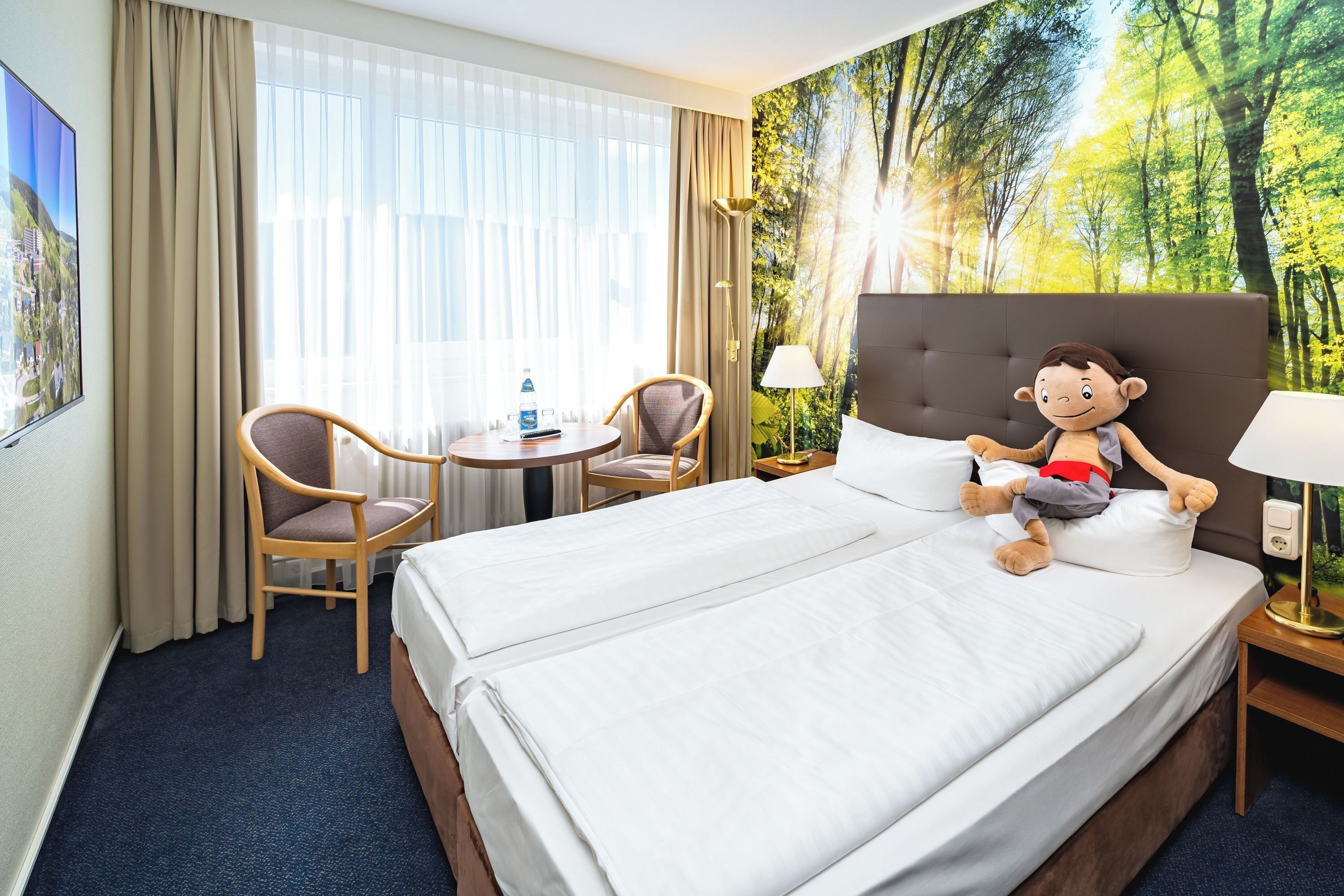 Een hotelkamer met twee bedden, een bureau, stoelen, een lamp en een muurschildering met een bosmotief.