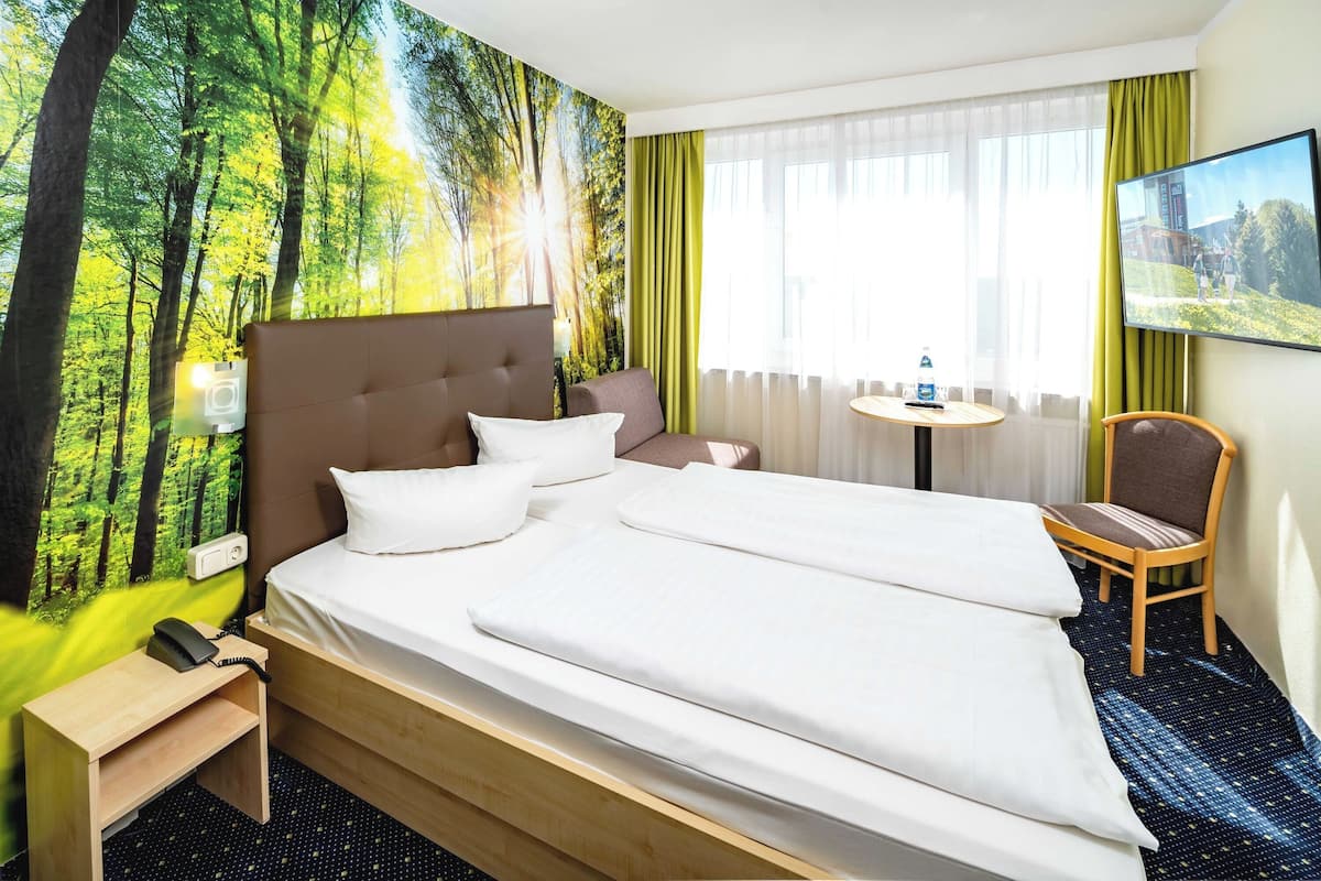 Une chambre d’hôtel avec un grand lit, une table de chevet avec un téléphone, une chaise, une petite table avec une bouteille et un téléviseur fixé au mur.