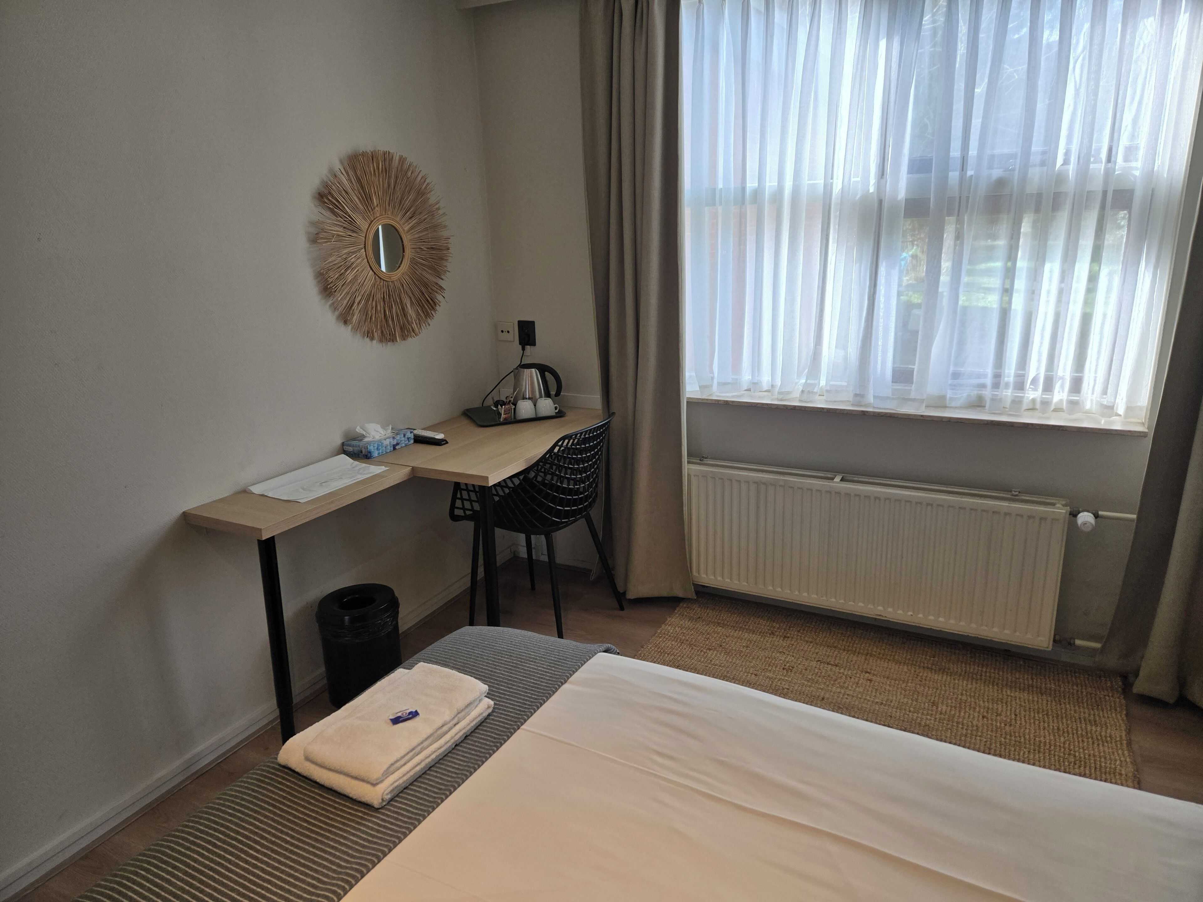 Una habitación de hotel con una cama, un escritorio con una silla, un radiador y una ventana con cortinas.