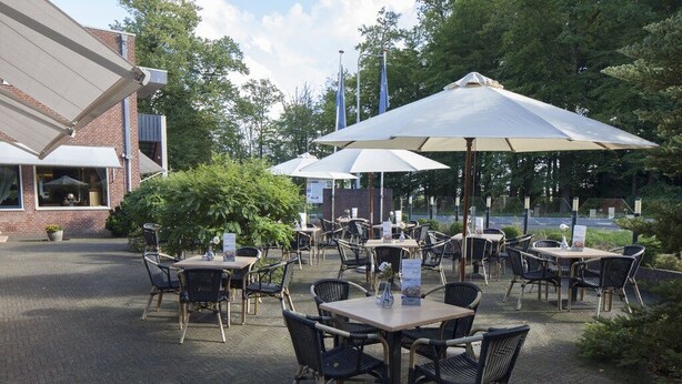 An outdoor patio area with tables and chairs, umbrellas, and a brick building in the background. - Hotel de Grote Zwaan (De Lutte)