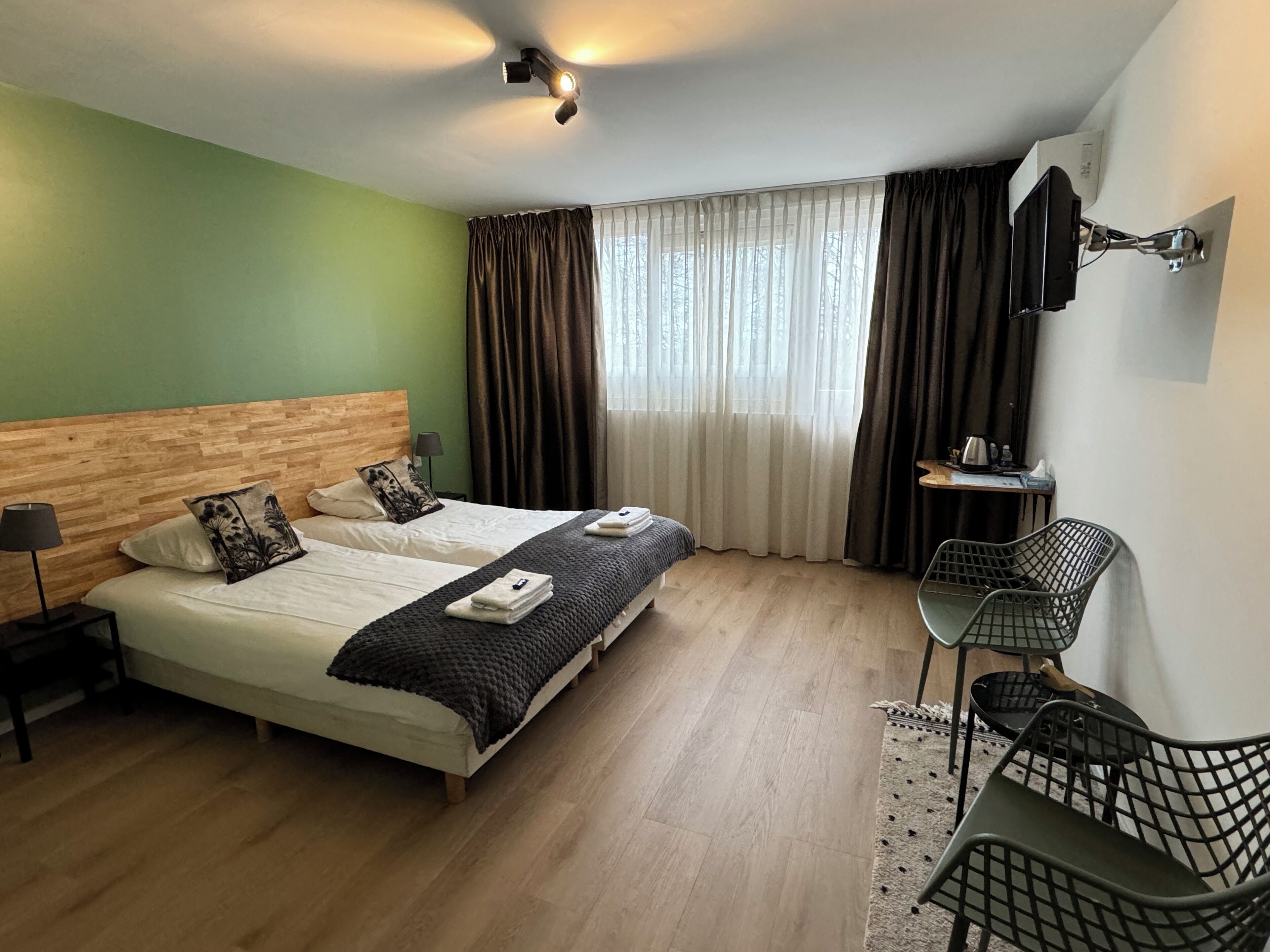 A hotel room with a wooden headboard bed, a desk with a chair, a window with curtains, and a ceiling light.