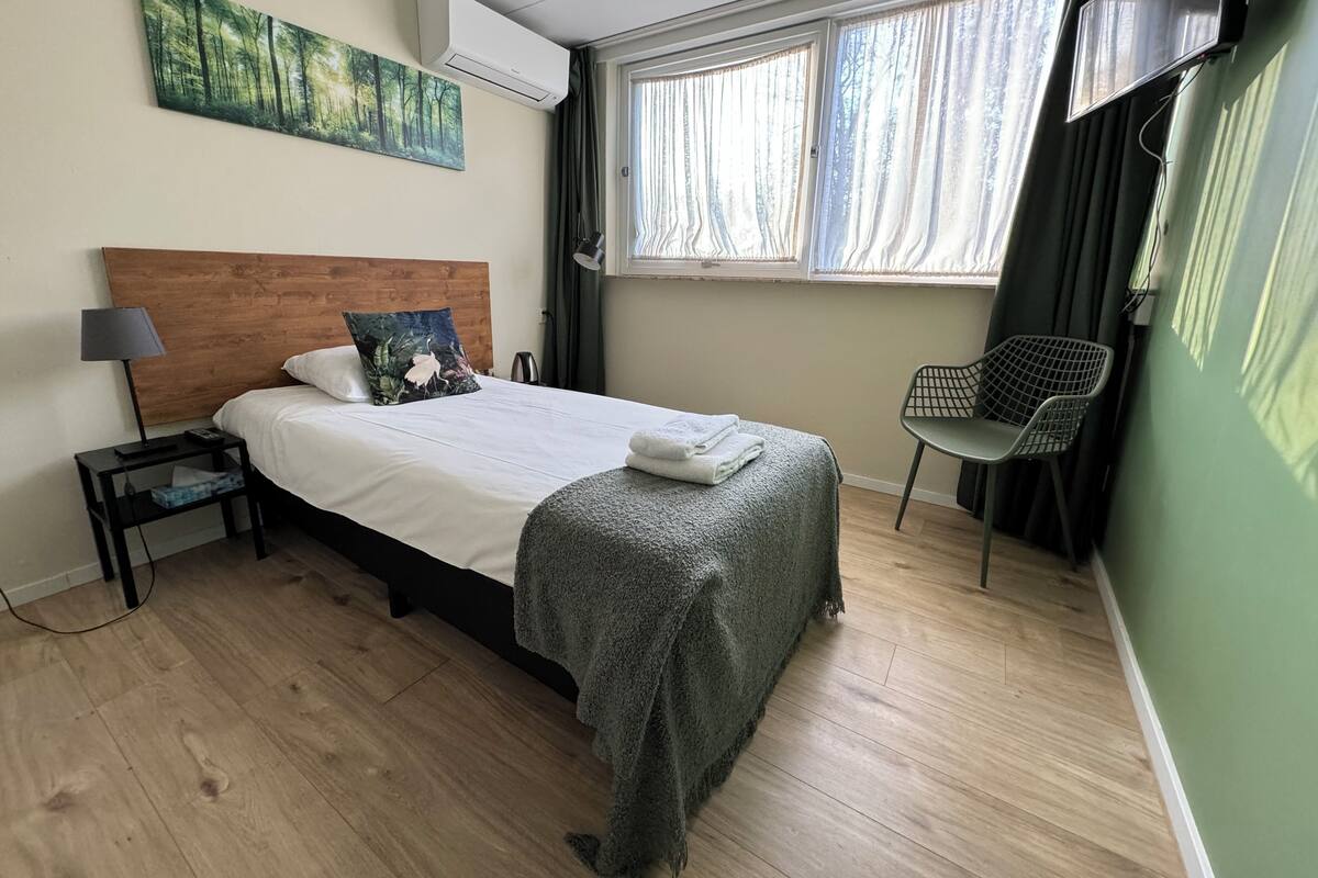 A neatly arranged hotel room with a wooden headboard bed, a bedside table with a lamp, a green chair, and a window with curtains.