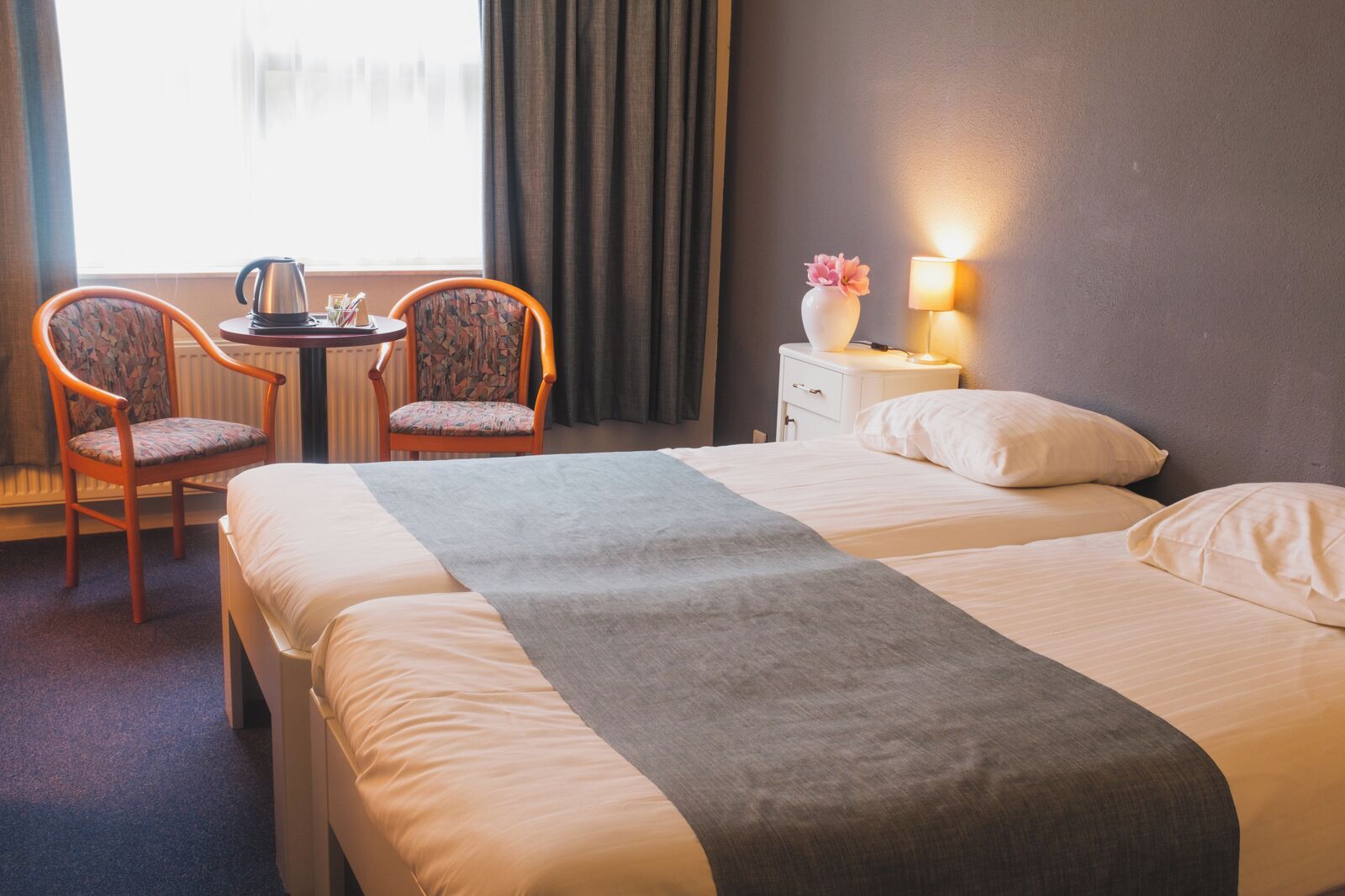 A hotel room with two beds, a small table with a kettle, two chairs, a nightstand with a lamp, and a window with curtains.