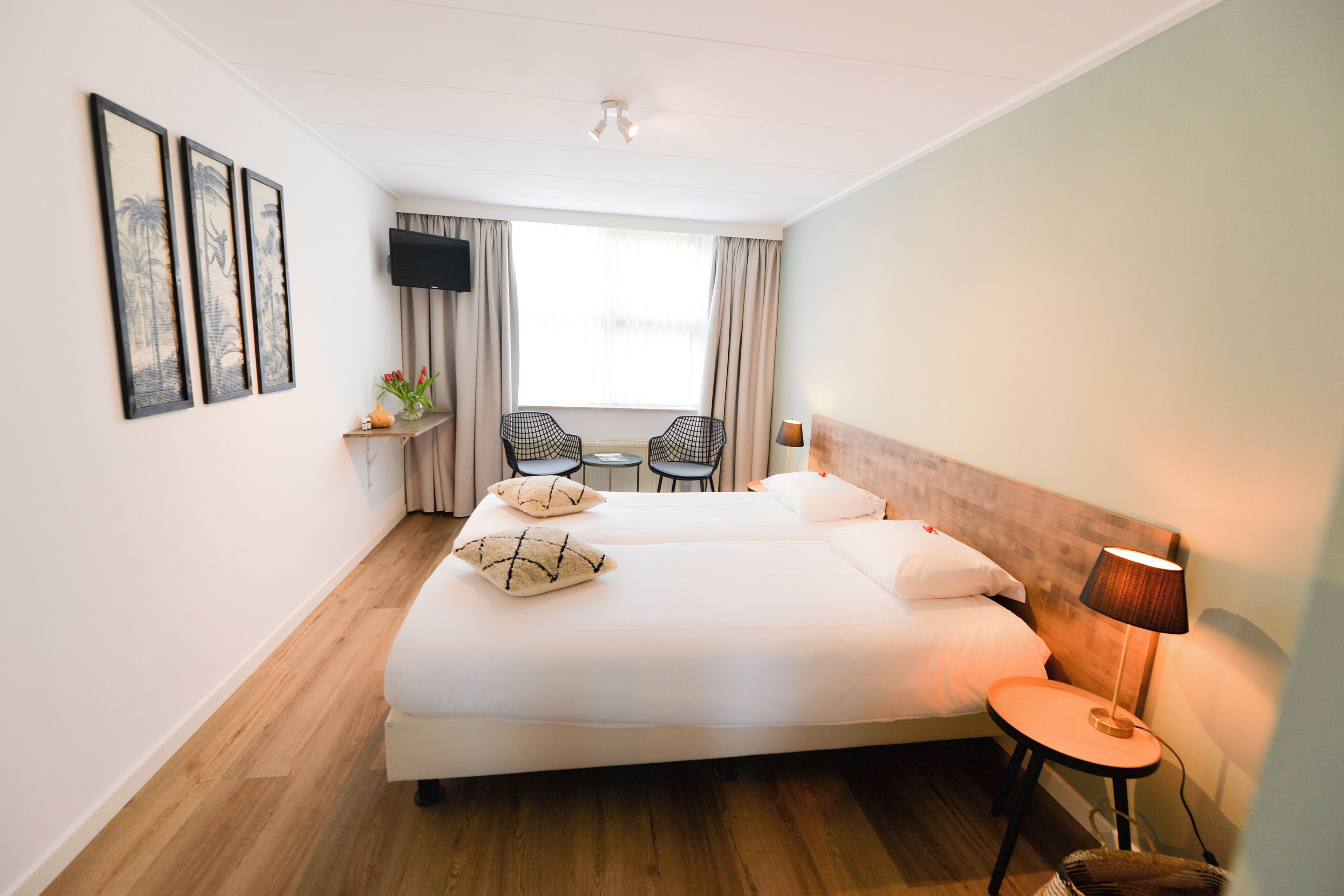 A hotel room with a large bed, a small table with chairs, a TV mounted on the wall, and a window with curtains.