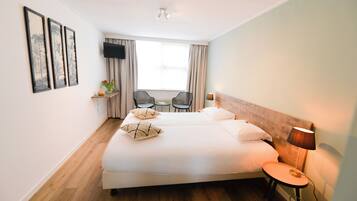 A hotel room with a large bed, a small table with chairs, a TV mounted on the wall, and a window with curtains.
