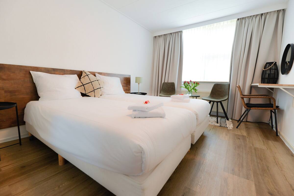 A hotel room with a large bed, two bedside tables, a chair, a desk, and a window with curtains.