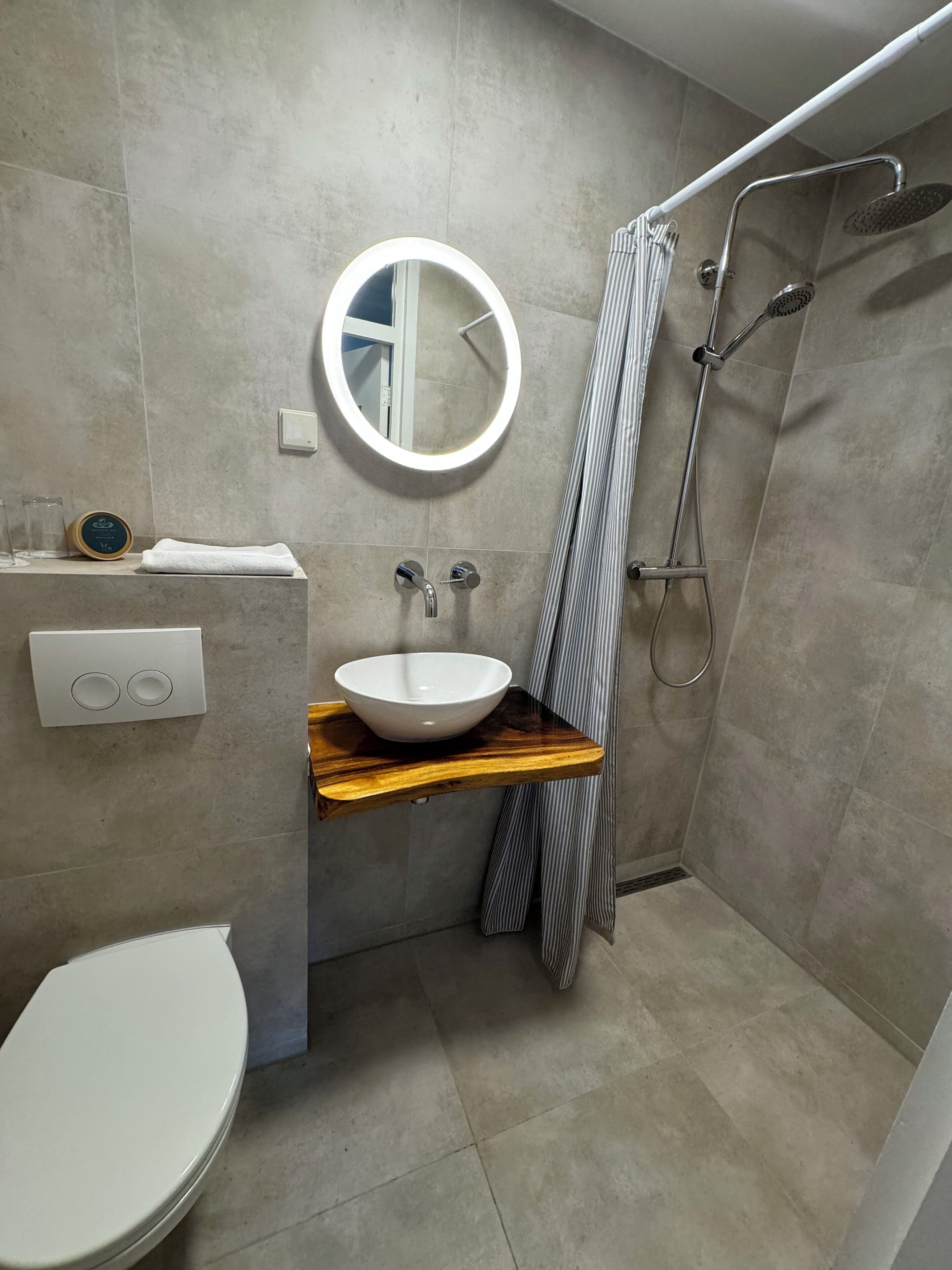A modern bathroom with a white toilet, a wooden vanity with a white basin, a round mirror with a light, and a shower area with a metal showerhead and curtain.