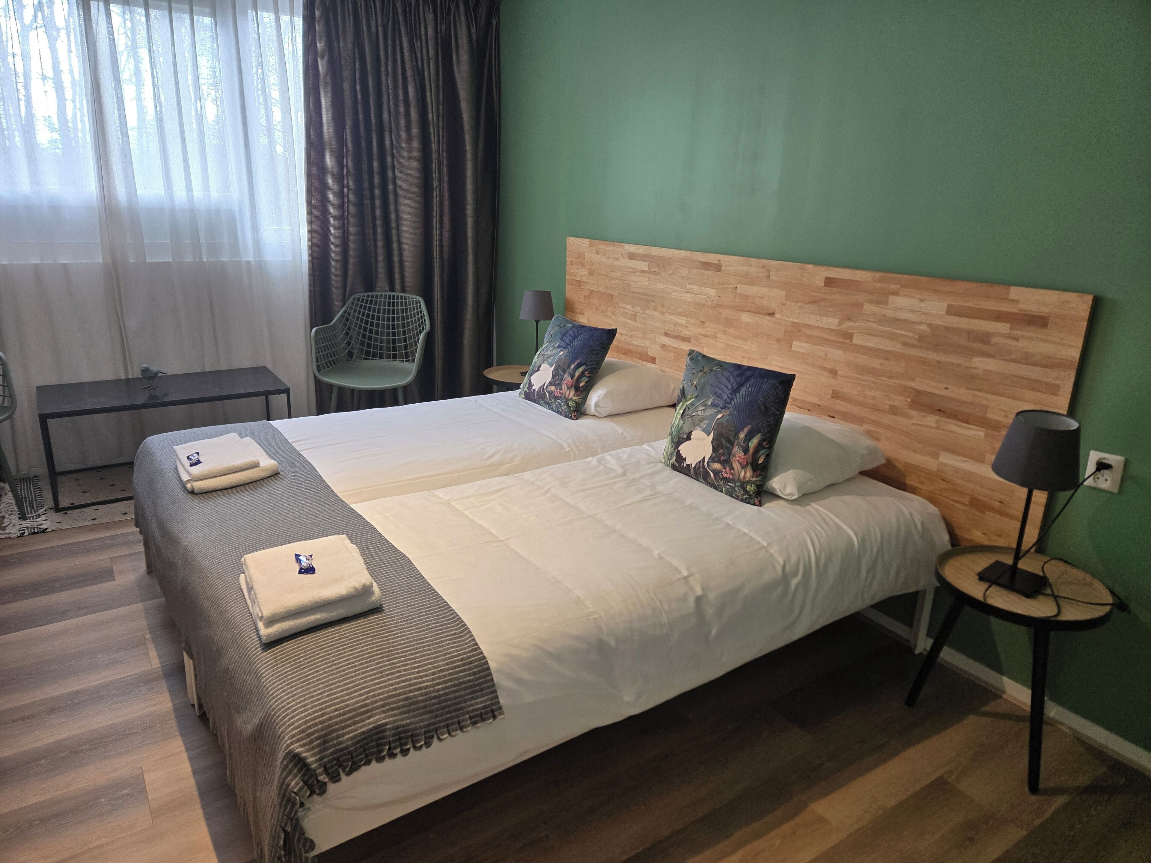 Una cama doble con cabecera de madera, dos almohadas con motivos florales, una mesita de noche con lámpara y una silla.