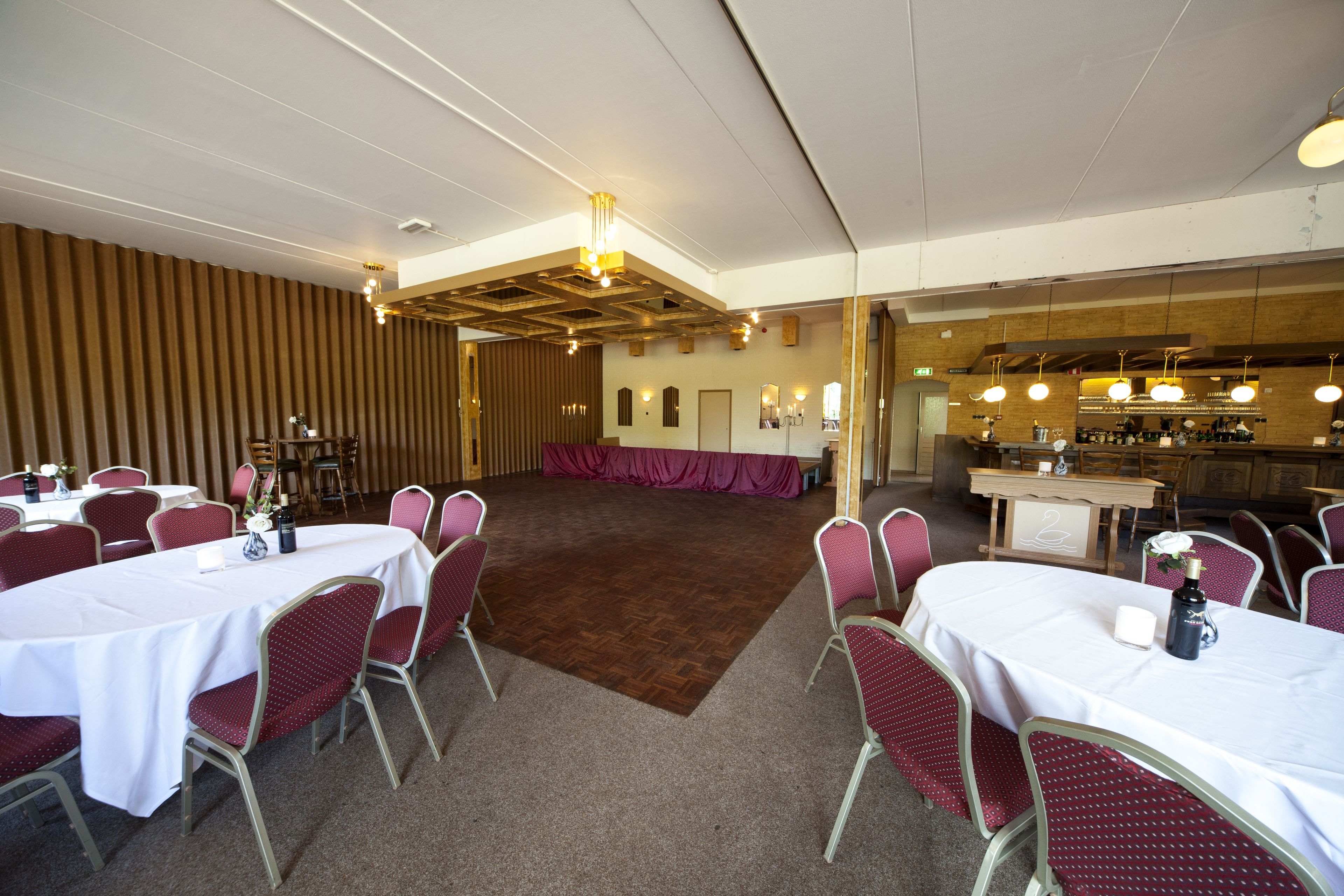 Een feestzaal met ronde tafels en stoelen, een houten plafond en een bar.