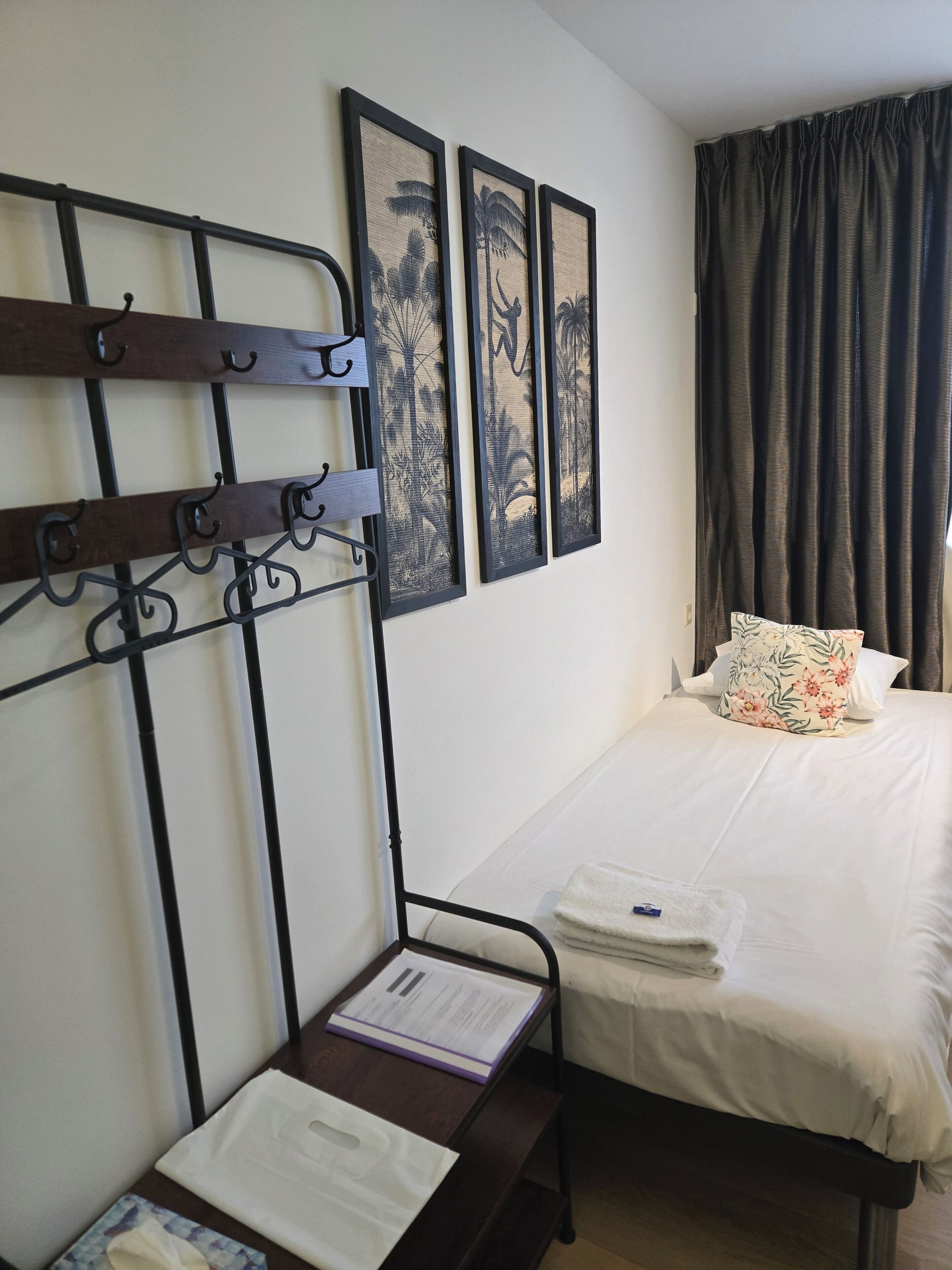 A hotel room with a bed, a bench with hangers, and three framed artworks on the wall.