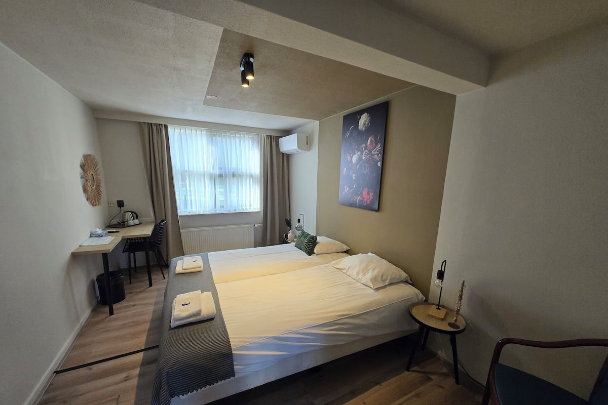 A hotel room with a bed, a desk, a chair, a small table, and a painting on the wall.