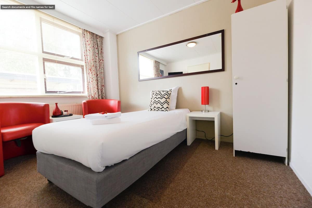 A hotel room with a bed, a red chair, a desk, a mirror, and a wardrobe.