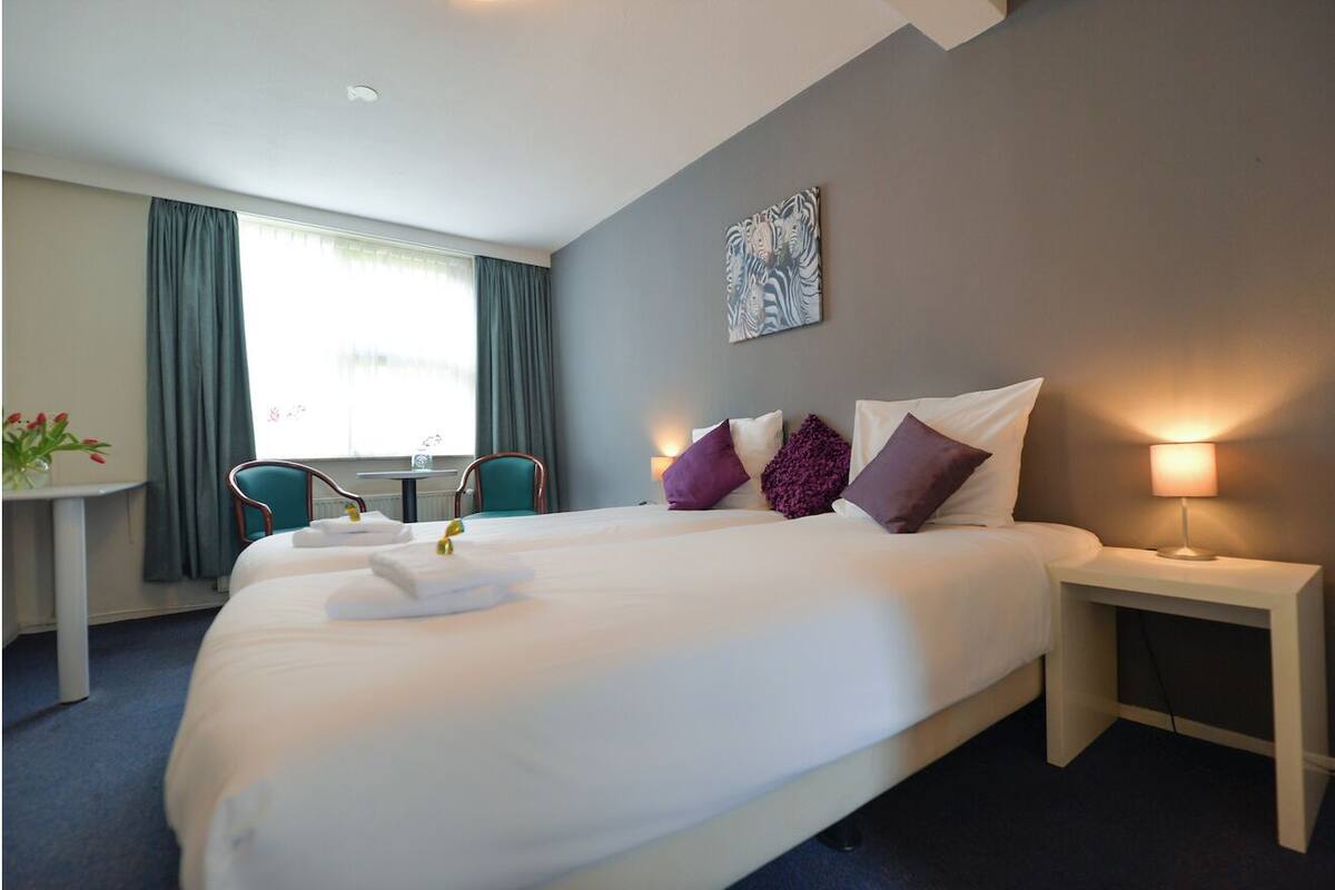 A hotel room with a large bed, a bedside table with a lamp, a chair, a small table with flowers, and a window with curtains.