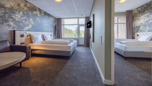 Ein Hotelzimmer mit zwei Betten, einem Sofa, einem Fernseher und einem großen Fenster mit Blick auf Bäume und eine Backsteinmauer.
