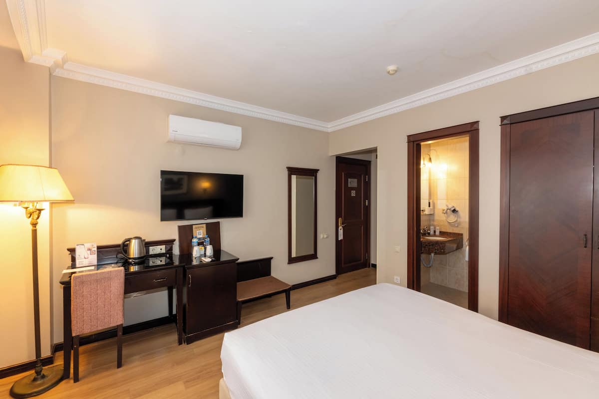 Quarto de hotel com cama, escrivaninha, cadeira, televisão e banheiro visível através de uma porta aberta.