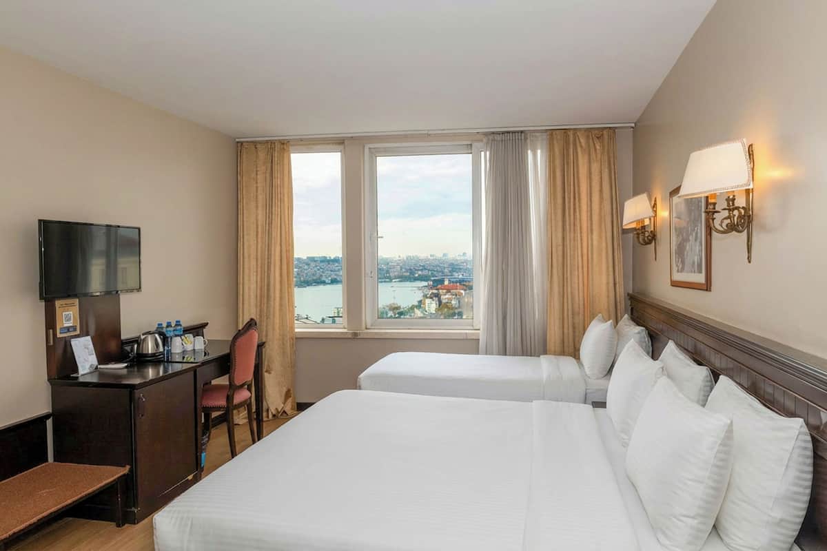 Quarto de hotel com duas camas, uma secretária com televisão e vista para a cidade através de grandes janelas, com uma vista urbana da janela.