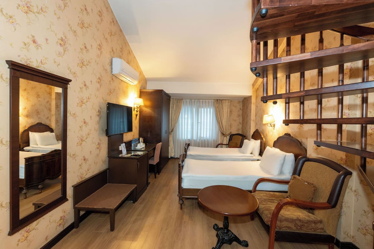 Quarto de hotel com cama, escrivaninha, cadeira, mesa pequena, espelho e escada.