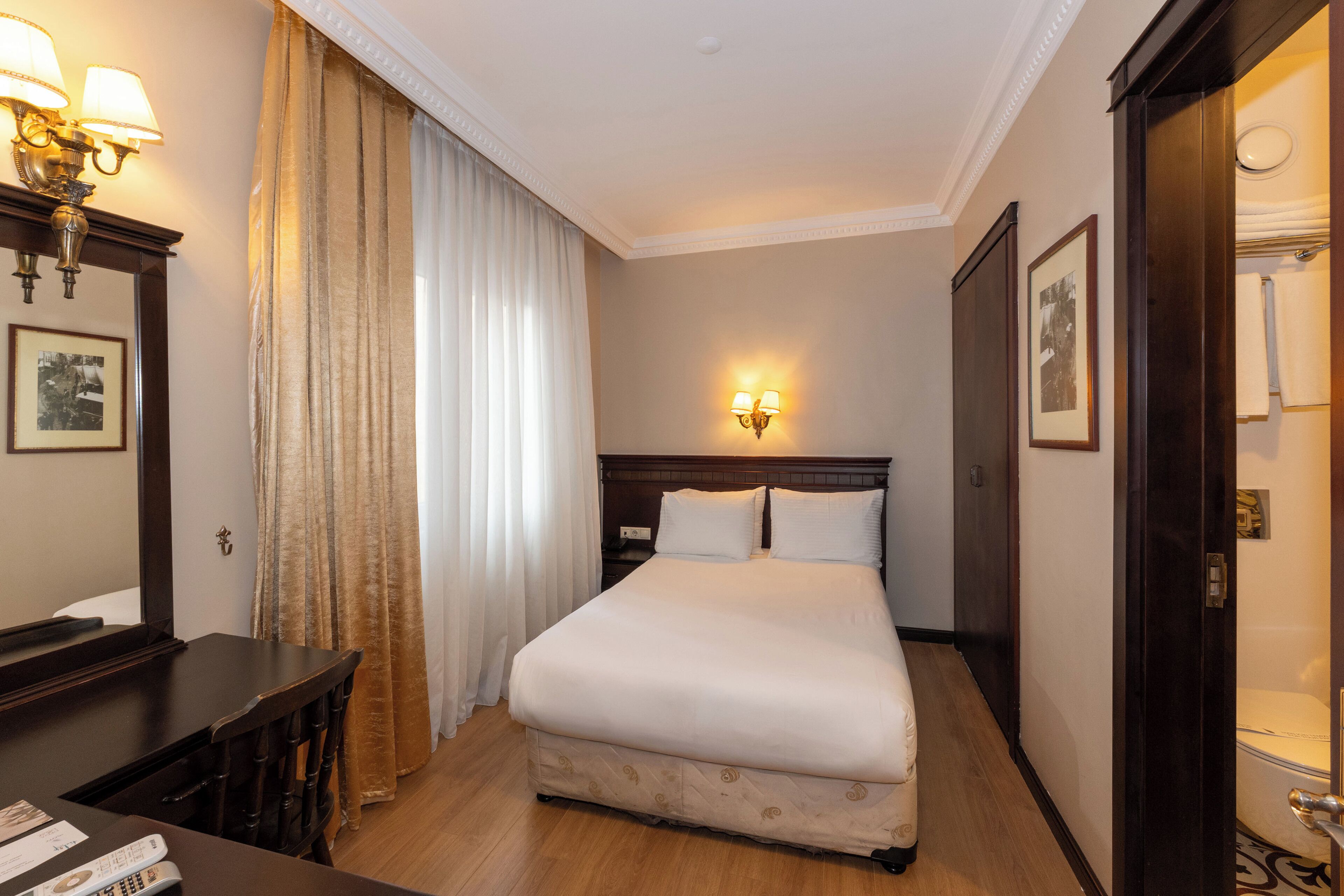 A hotel room with a large bed, a bedside table with lamps, a mirror, and a door leading to a bathroom.