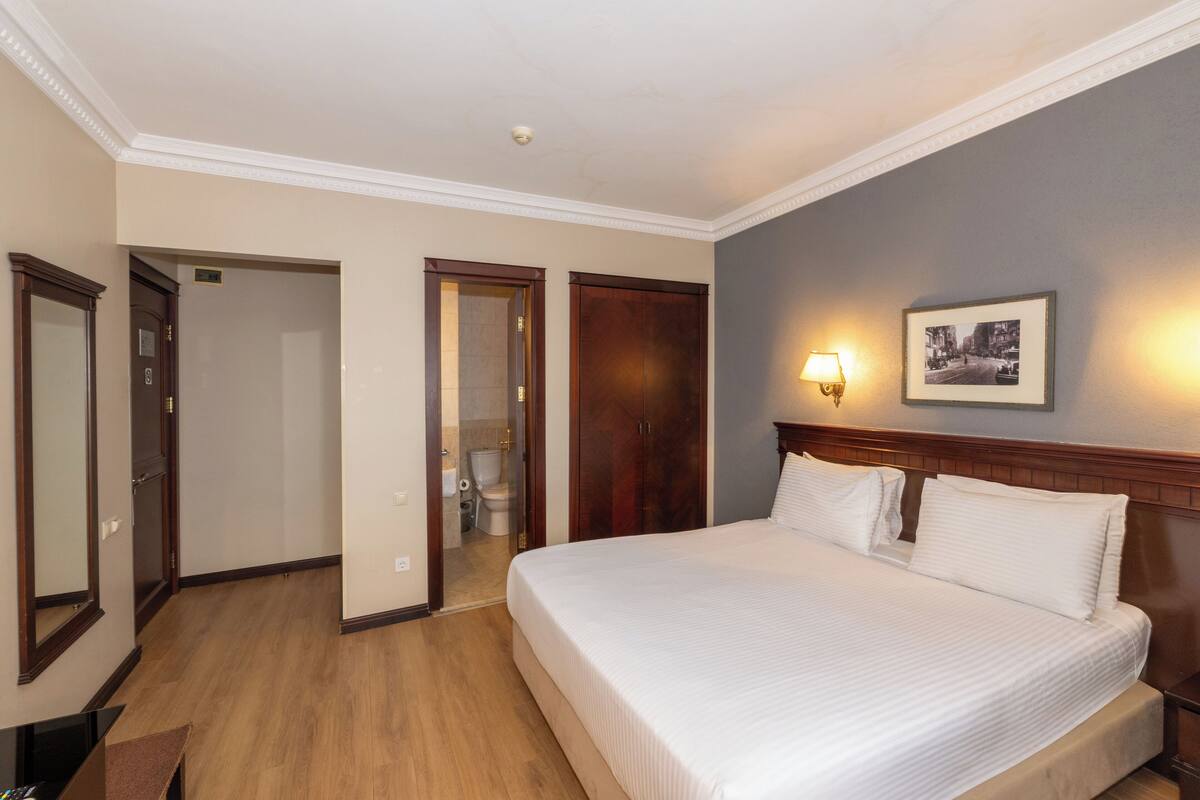 Quarto de hotel com uma cama grande, cabeceira de madeira, um quadro na parede e um banheiro visível através de uma porta aberta.