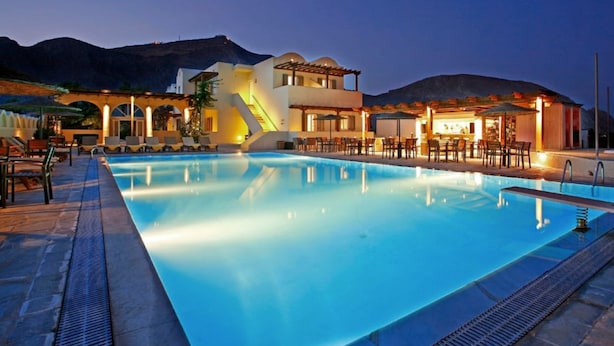 A swimming pool with lounge chairs and a restaurant area, surrounded by mountains. - Thera Mare (Santorini)