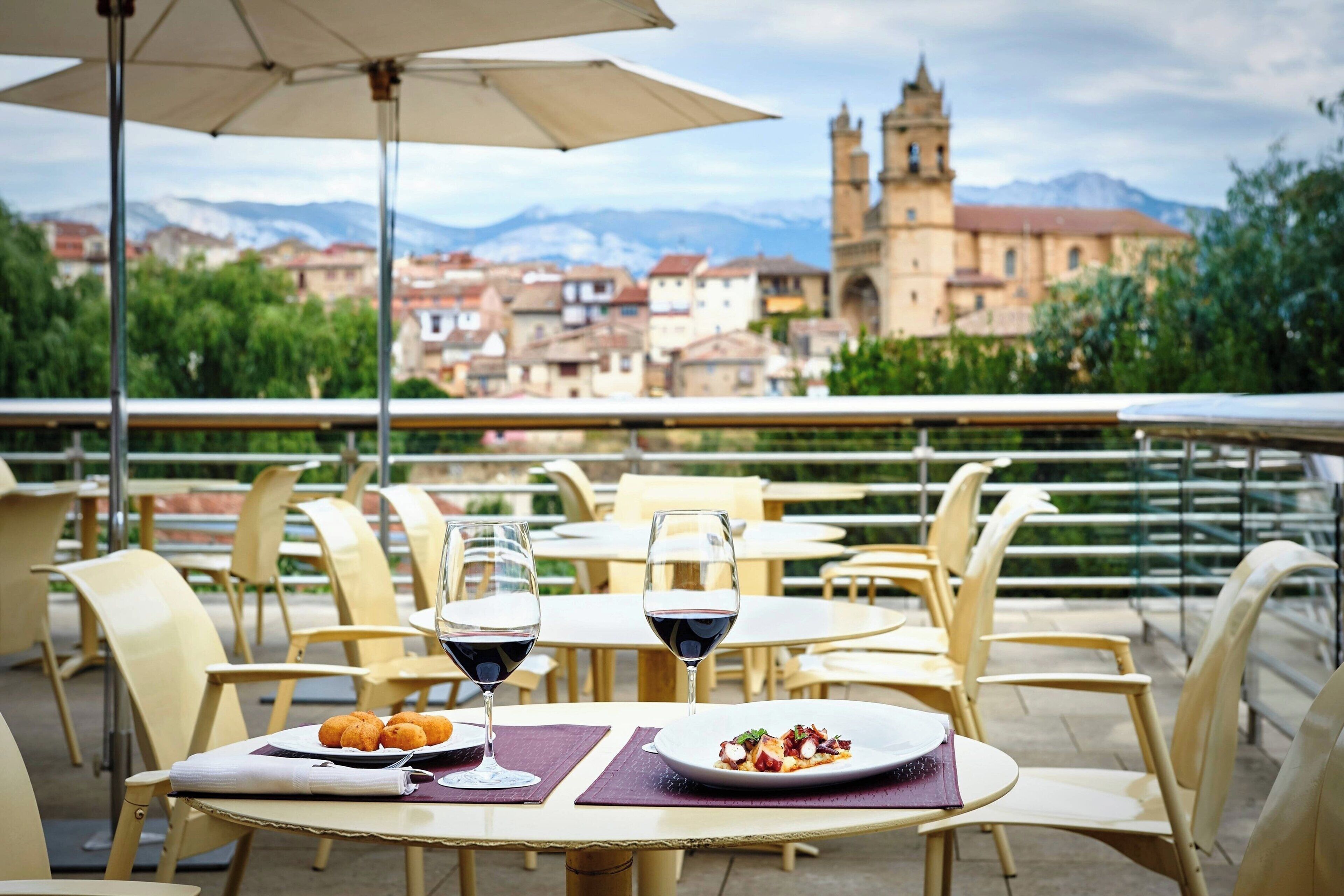 An outdoor dining area with tables and chairs, a table with a plate of food and two wine glasses, and a view of a historic building in the background.