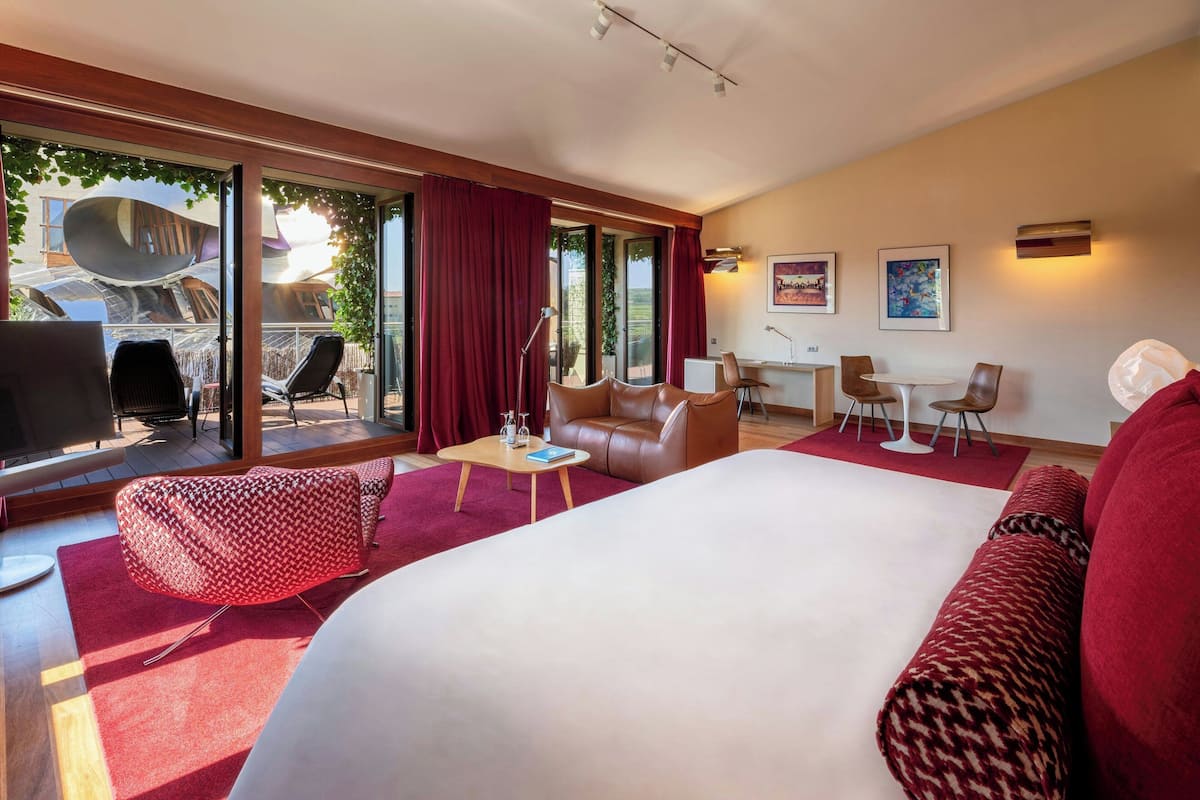 A modern hotel room with a large bed, a seating area with a sofa and chairs, a small table, and a view of the outdoors through large windows.