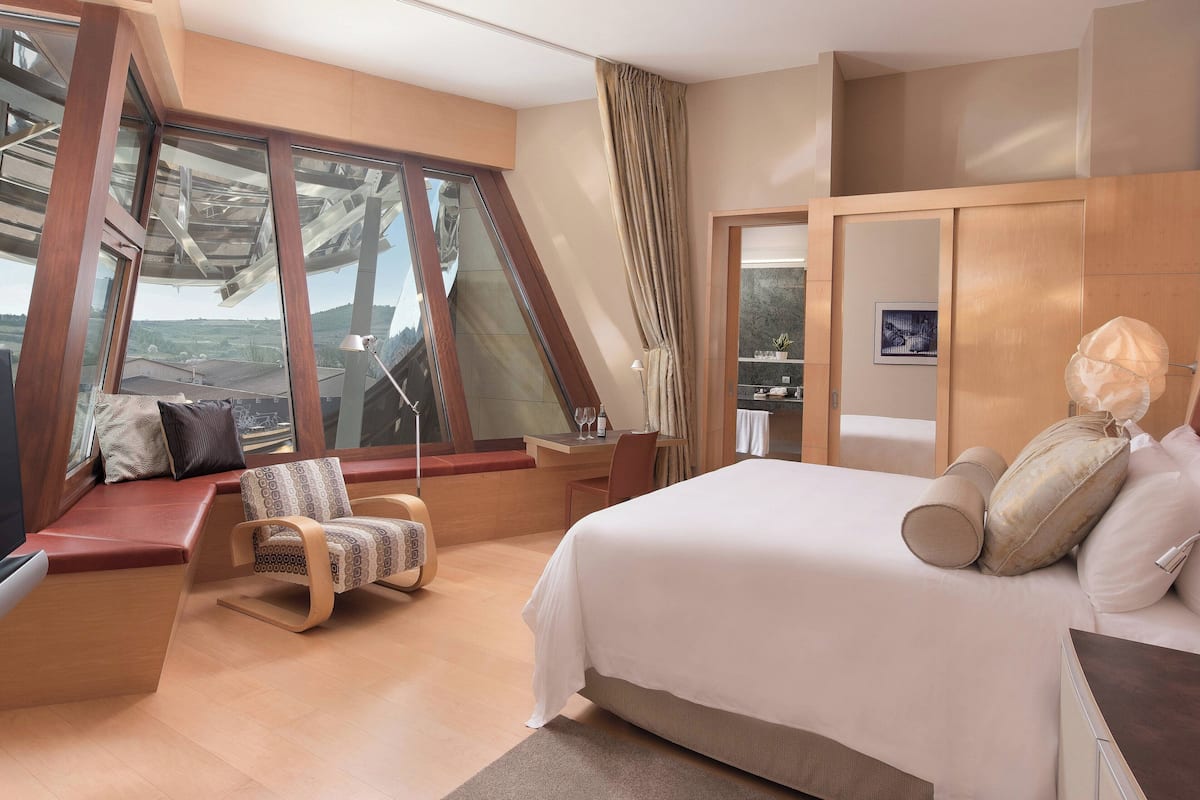 A modern hotel room with a large bed, a seating area with a chair, a desk, and a view through large windows.