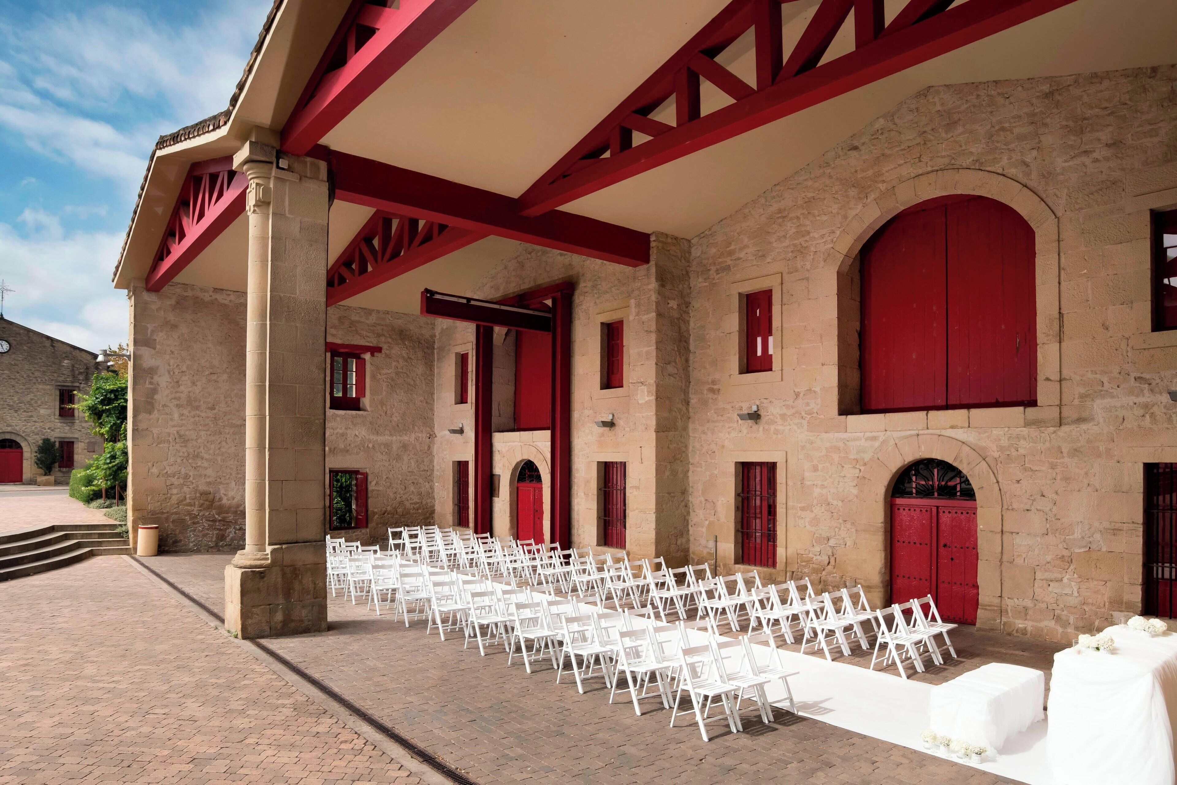 A building with red-framed windows and doors, white chairs arranged in rows, and a brick-paved courtyard.