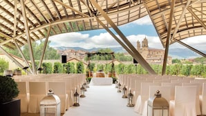 Un espai per a cerimònies nupcials a l'aire lliure amb files de cadires blanques i un passadís blanc, un sostre cobert i vista a un edifici històric al fons.