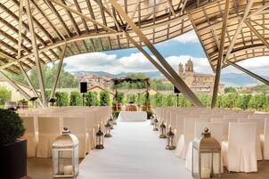 Espaço para casamento ao ar livre com fileiras de cadeiras brancas e um corredor branco, uma cobertura acima e vista para um edifício histórico ao fundo.