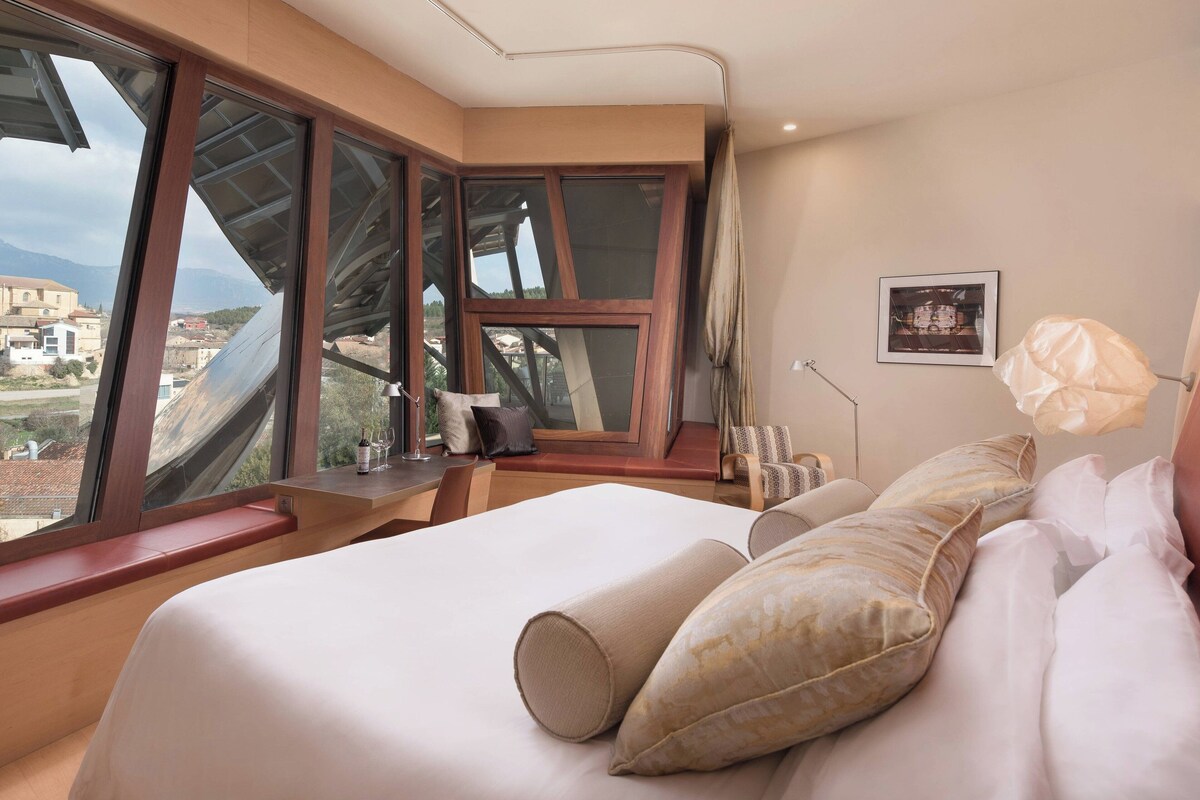 Une chambre d’hôtel avec un grand lit, plusieurs fenêtres, une table de chevet, une lampe et un tableau encadré au mur.