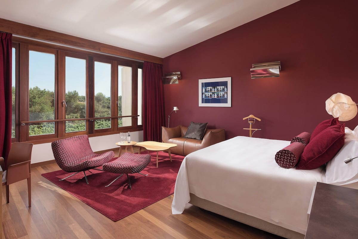 A modern hotel room with a large bed, a seating area with a table, and a view of greenery through large windows.