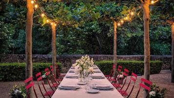 An outdoor dining area with a long table set for a meal, surrounded by red chairs, under a canopy of trees with string lights.