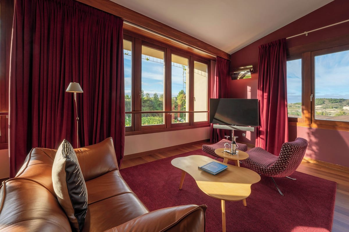 A modern living room with a leather sofa, a patterned armchair, a small round table, and a flat-screen TV. Large windows with red curtains provide a view of the outdoors.