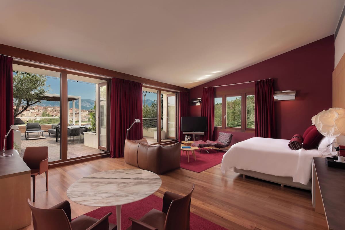 A modern hotel room with a large sliding glass door leading to an outdoor terrace, a king-sized bed, a seating area with a TV, and a dining table.