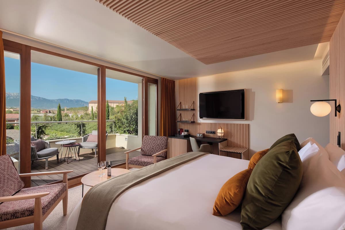 A hotel room with a large bed, a desk, a TV, and a balcony with outdoor seating.