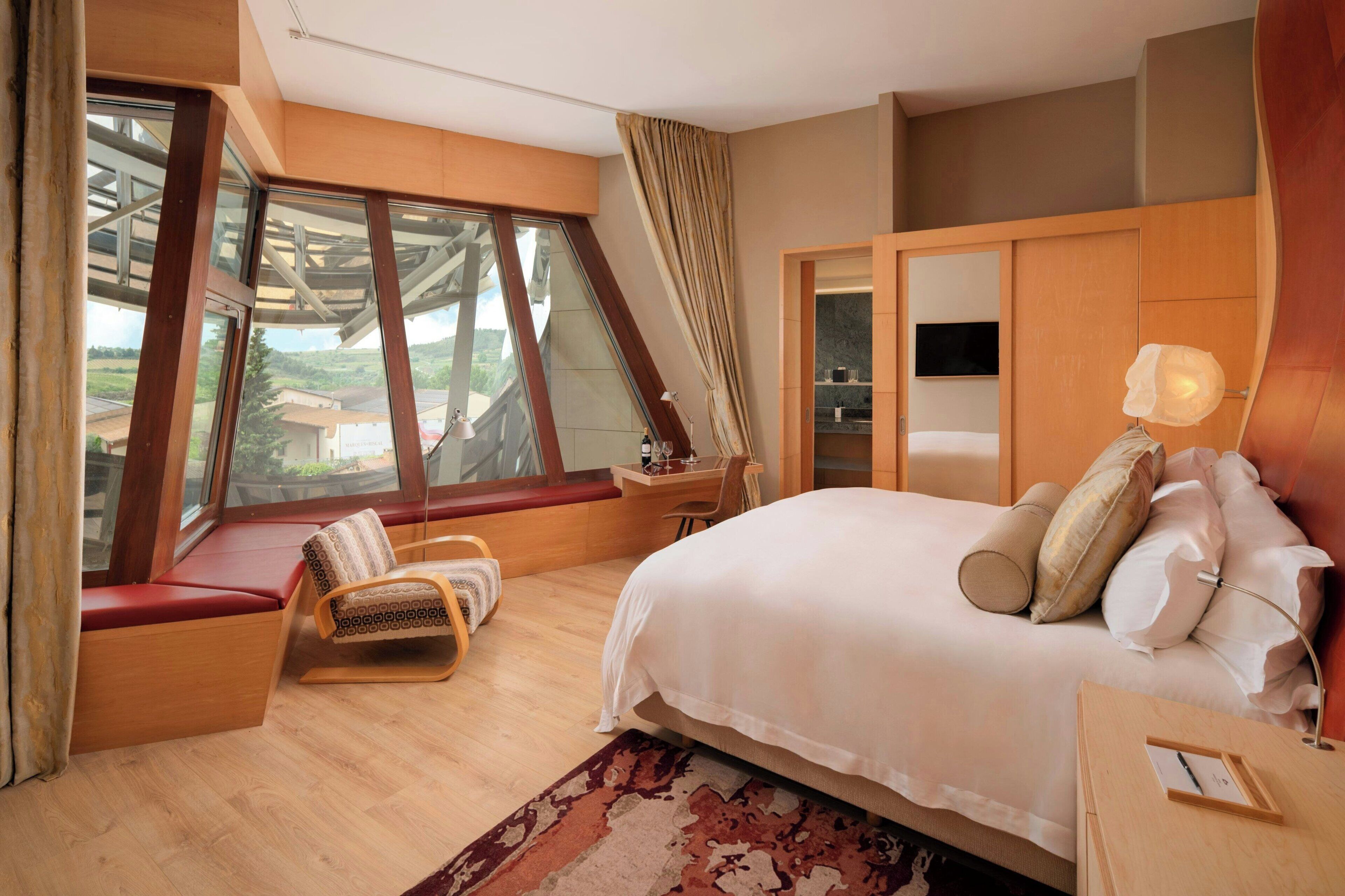 Un dormitorio con una cama grande, un banco junto a la ventana, un escritorio y televisión.