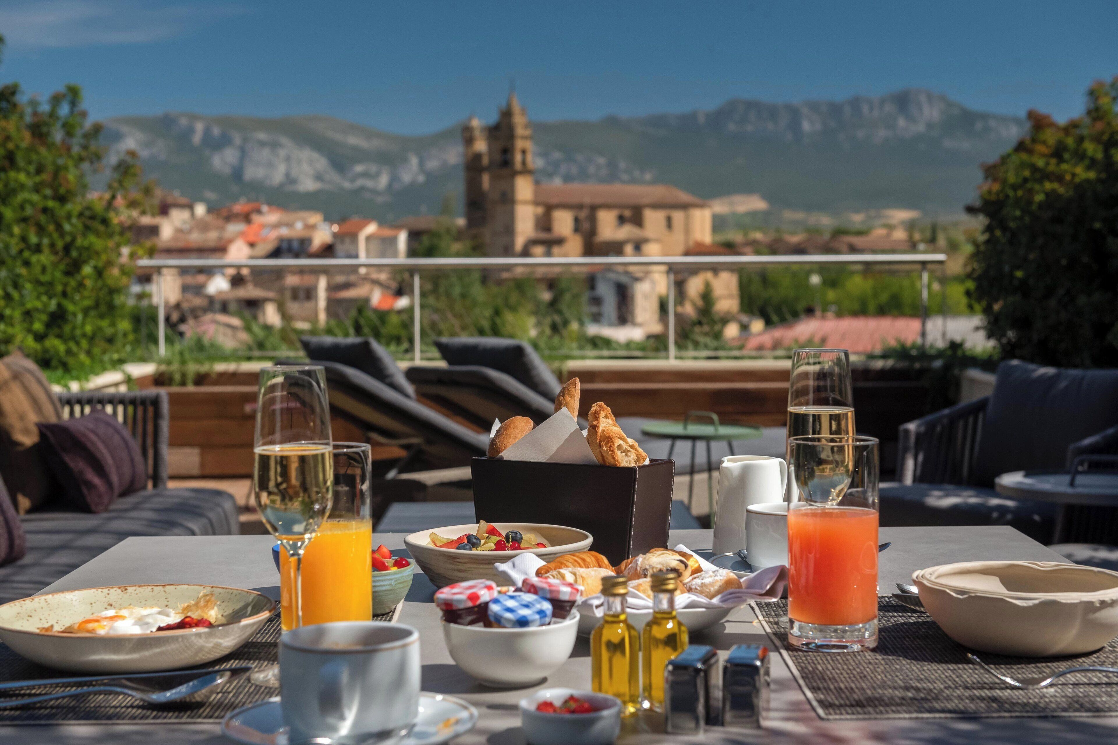 Mesa de pequeno-almoço com diferentes alimentos e bebidas, com vista para uma aldeia pitoresca e montanhas.