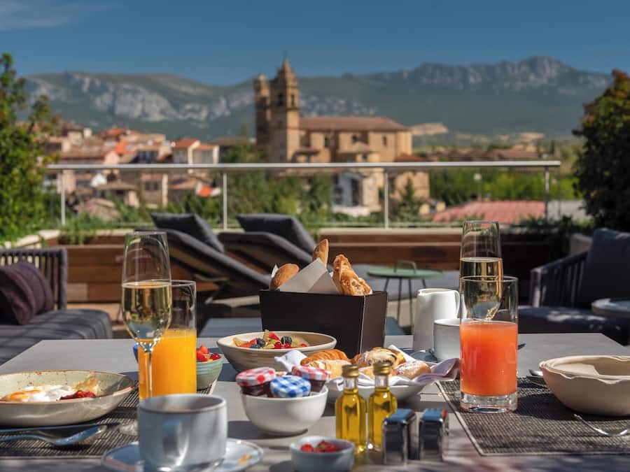 Mesa de pequeno-almoço com diferentes alimentos e bebidas, com vista para uma aldeia pitoresca e montanhas.