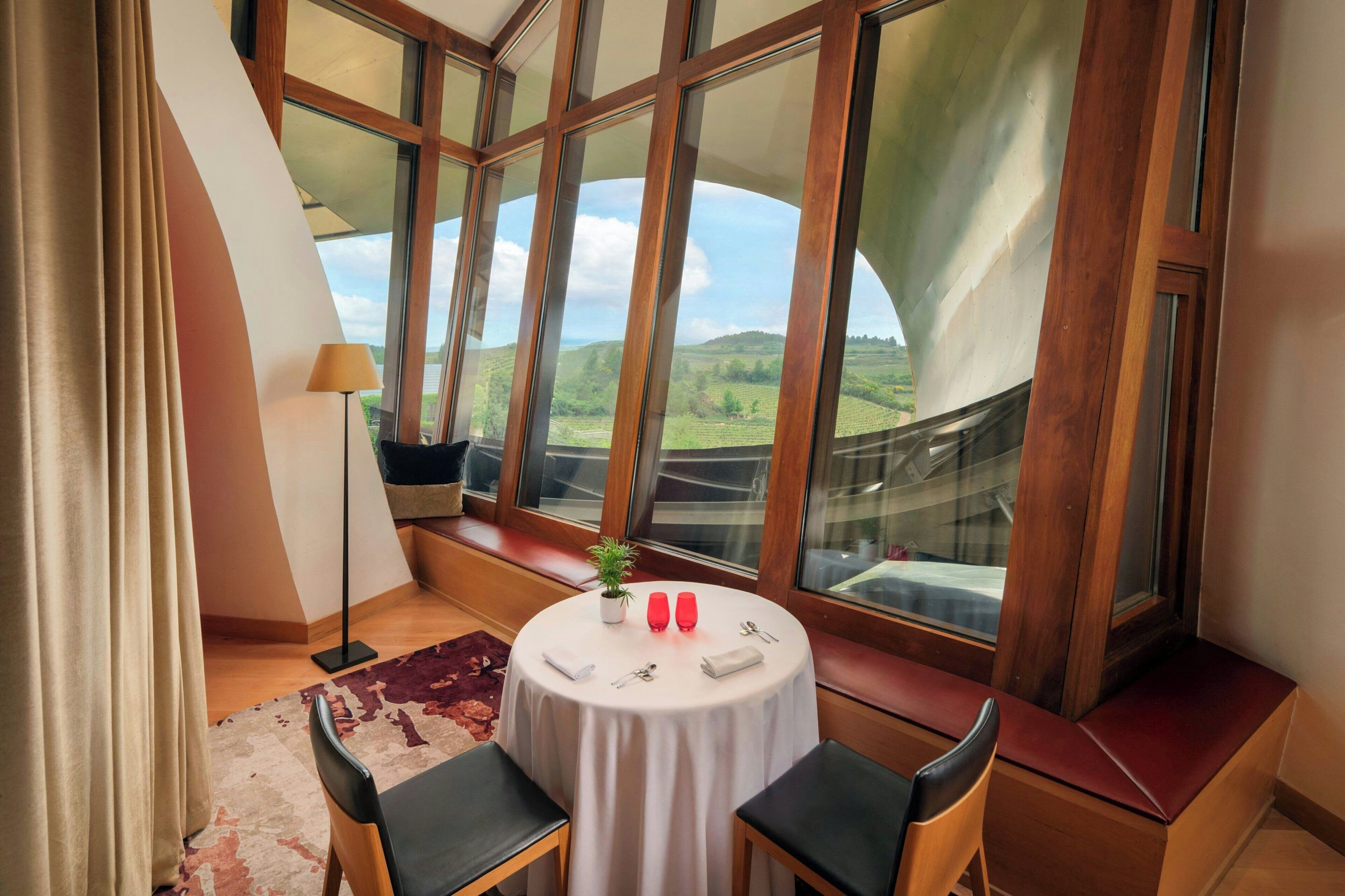 A room with a round table, two chairs, a potted plant, and a view of green hills through large windows.